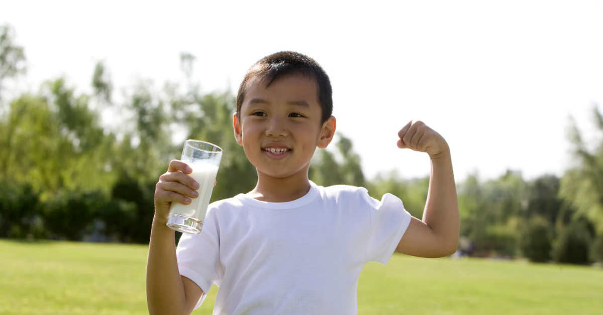 Lächelnder Junge hält ein Glas Milch und zeigt seinen angespannten Oberarm – Calcium Dosierung bei Kindern für Knochen und Muskeln.